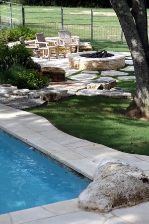 Edge of a pool with stone tiles and a fire pit with chairs in the distance