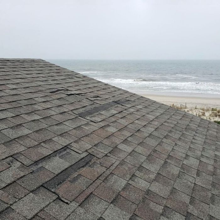 Strong coastal winds tear and lift shingles on a residential roof.