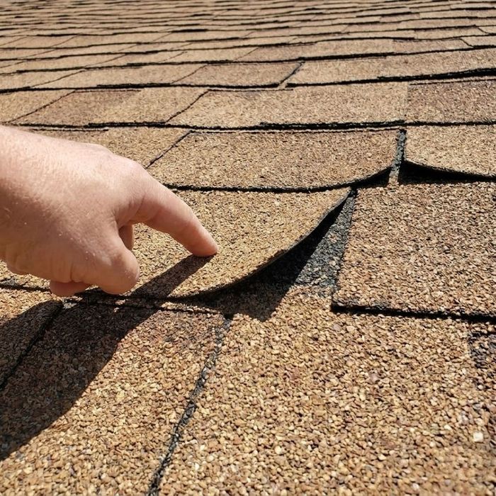A close-up photograph of a hand pointing to brittle, sun-damaged roofing shingles that are curling at the edges.
