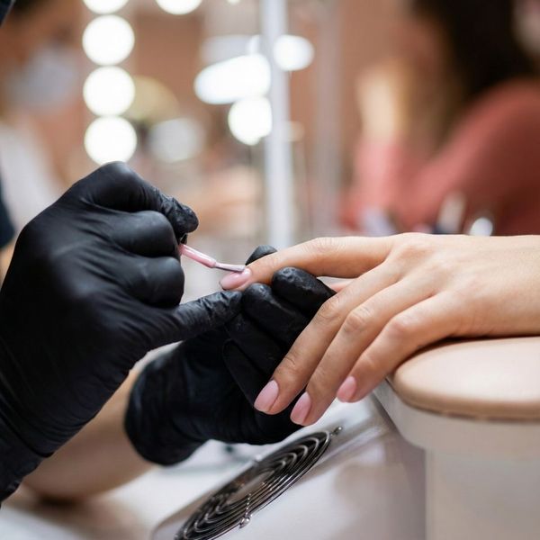 A close-up of a technician's hands in black gloves carefully painting a client's nails with a pink polish, with a warm, blurred background suggesting a friendly salon environment.
