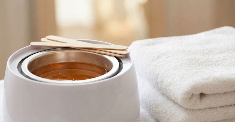 Professional white wax warmer filled with melted amber hard wax and wooden spatulas, next to a stack of clean white towels on a table.