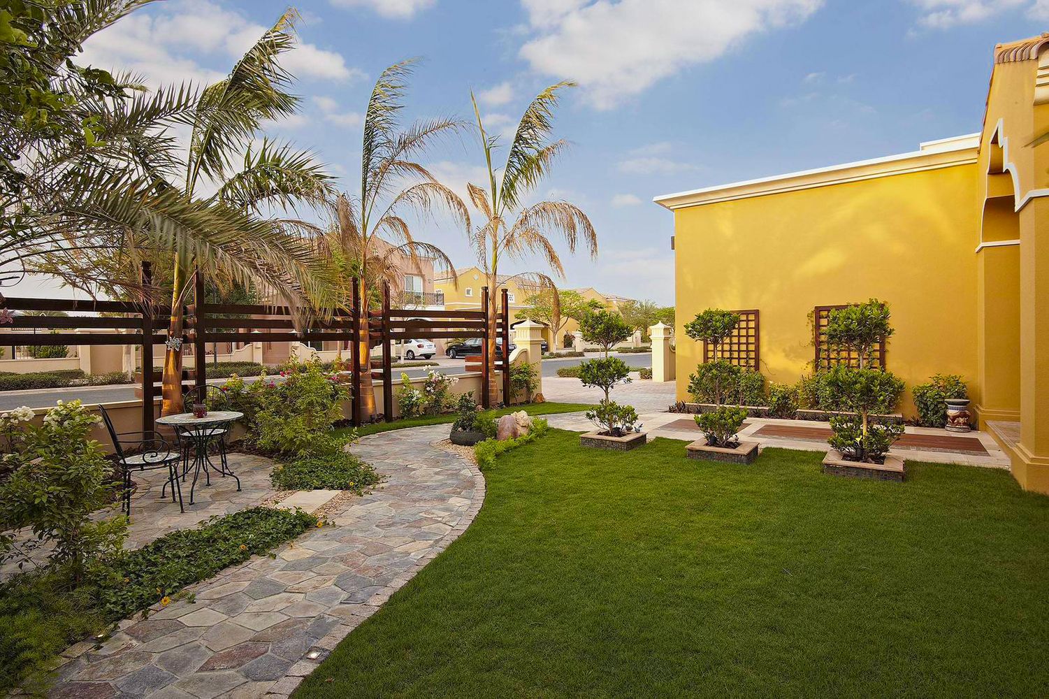 a big house with artificial grass and a lot of plants surrounding, also a chair and a table near the palm trees with a paver walkway 