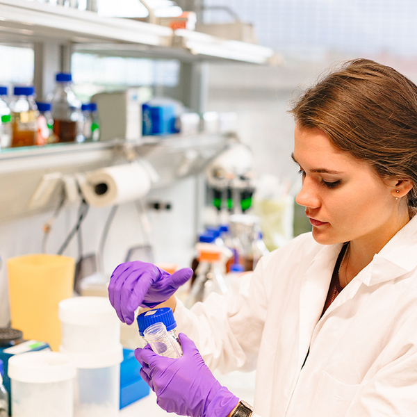 lab technician unscrewing plastic bottle, with chemicals in the background