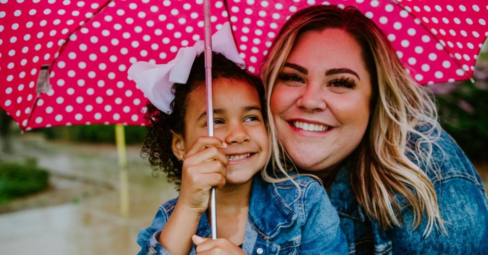 mother and daughter smiling underneath a pink umbrella on a rainy day