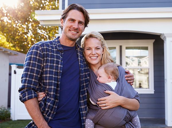 Couple standing in front of their home smiling and holding their baby