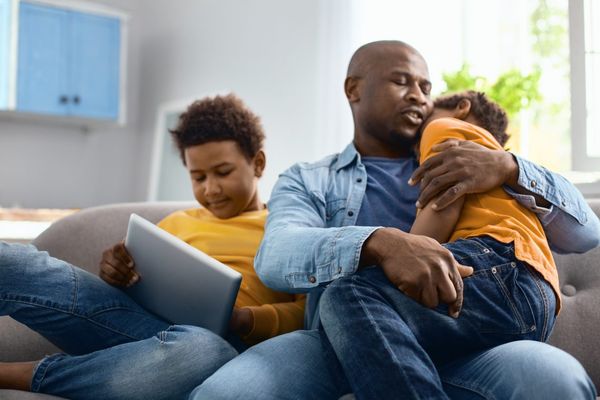 father consoling upset child while other child sits next to them on tablet