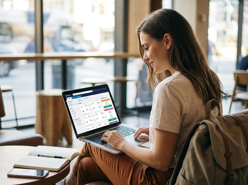 A focused young woman uses a laptop in a bright cafe, reviewing flight options on a travel booking website. A backpack, notebook, and phone are nearby. Young Woman Planning Travel on Laptop in Cafe