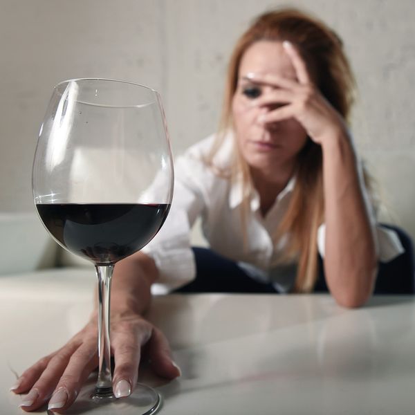Drunk woman with one hand covering her face and the other reaching for a glass of wine.