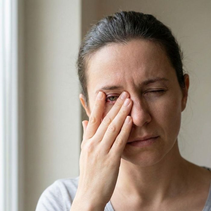 A close-up of a woman with a pained expression, holding her hand over her reddened eye.