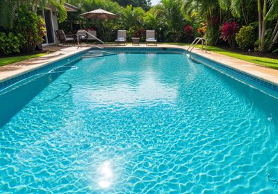 A rectangular swimming pool filled with sparkling turquoise water sits amidst lush tropical foliage. Two lounge chairs with rolled towels face the pool, shaded by a large umbrella. Palm trees and colorful flowers add to the tranquil, resort-like ambiance. A ladder provides access to the inviting, sun-drenched water, perfect for a refreshing swim. Swimming pool with turquoise water and surrounding tropical foliage