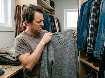 A man with a concerned expression holds up a gray, knitted sweater with multiple holes, suggesting moth damage, in a cluttered closet. Man holding a damaged, moth-eaten sweater in a closet