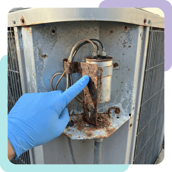 A close-up photograph of an HVAC technician's gloved hand pointing at a corroded electrical capacitor bracket inside the open service panel of an outdoor air conditioning condenser unit. The metal casing of the unit shows signs of slight salt-air weatherin
