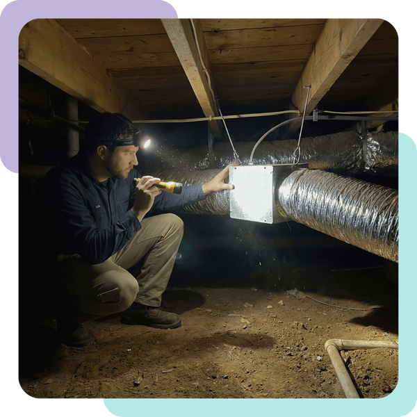 A photograph of an HVAC technician crouching in a dimly lit residential crawlspace, holding a bright flashlight. The beam is focused on a junction of silver flexible air ducting suspended from wooden floor joists, as the technician inspects it for leaks or