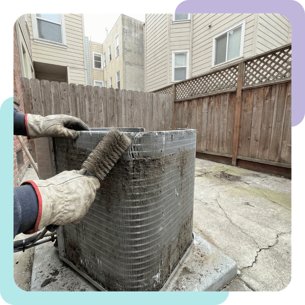 A close-up photograph of an HVAC technician's gloved hands using a stiff coil brush to clean accumulated dust and urban grime off the aluminum fins of a compact outdoor air conditioning condenser unit. The unit is situated on a small concrete patio in a de