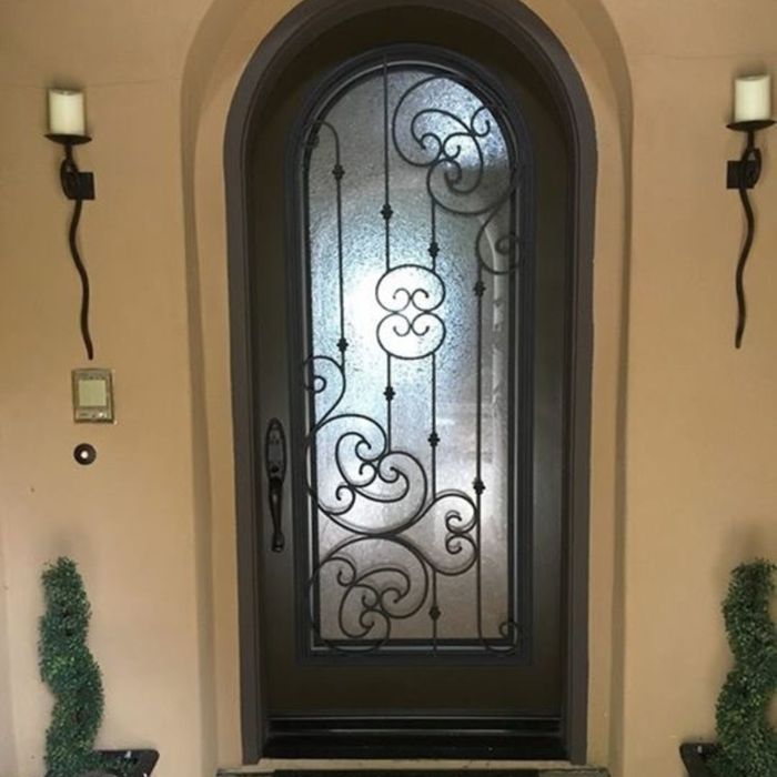 A dark arched door with an elegant wrought iron heart and scroll design over frosted privacy glass, flanked by wall-mounted candle sconces.