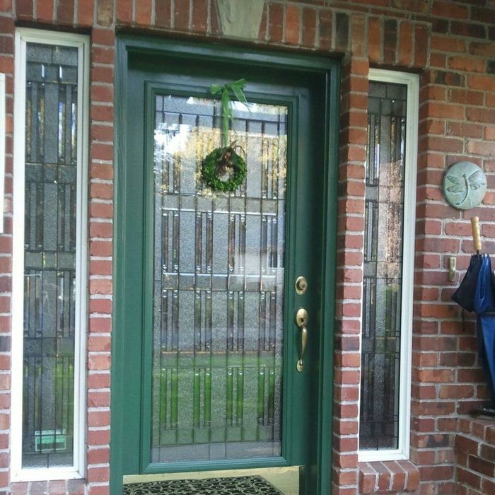 A forest green front door with a rectangular textured glass insert and a small evergreen wreath, set in a classic red brick wall with sidelights.