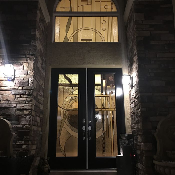 A night view of a modern black double-door entryway with geometric glass patterns illuminated from within, set between lighted stone pillars.