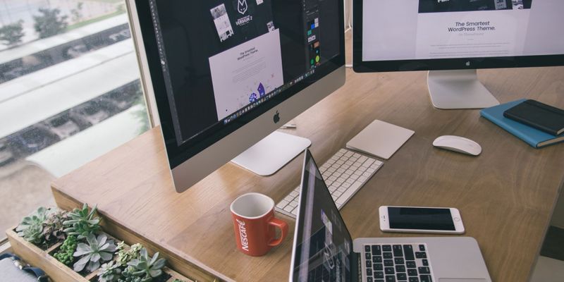 A high-angle view of a modern wooden office desk featuring two monitors, a laptop, a smartphone, and a red coffee mug.
