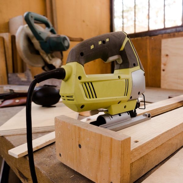 A green jigsaw sits on a wooden workbench in a workshop with its power cord visible.