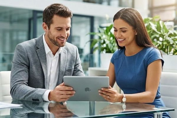 A man in a gray blazer and a woman in a blue dress sit at a glass-topped table in a bright modern office and smile at the business data graph with an upward arrow displayed on the tablet they both hold.
