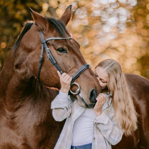 A woman affectionately leaning her head against a brown horse in a soft-focus outdoor setting.