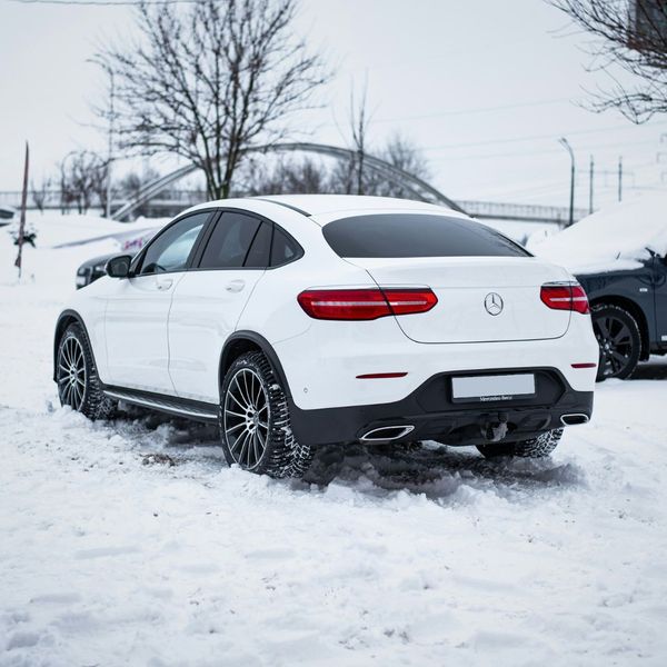 A white Mercedes-Benz SUV is parked in a snow-covered area, with its rear facing the camera. The car's tires are covered in snow, suggesting cold weather conditions.