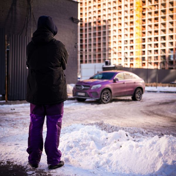 A person wearing a black jacket and purple pants stands in a snowy parking lot, with their back to the camera. A purple Mercedes-Benz SUV is in the background, partially obscured by snow piles.