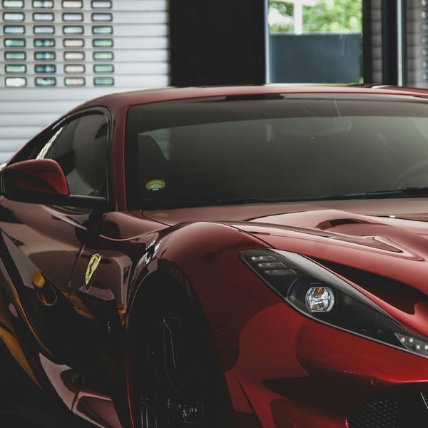 A detailed front quarter view of a sleek, red Ferrari sports car, with a focus on its sharp headlights and the iconic prancing horse logo. The background is slightly blurred, showing a modern interior space, suggesting a professional garage or showroom.