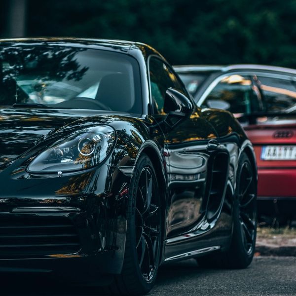 A close-up side view of a glossy black Porsche sports car. The reflection on the car's paint highlights its clean, polished surface. Another car is partially visible in the blurred background.