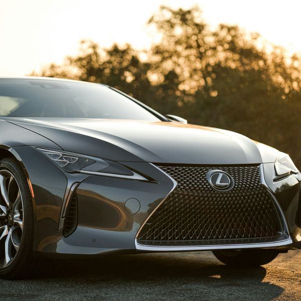 The front of a luxurious dark gray Lexus coupe, set against a backdrop of golden hour sunlight filtering through trees. The image emphasizes the car's intricate grille design and flawless exterior.