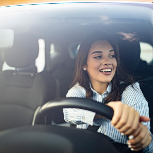 A smiling young woman with dark hair is in the driver's seat of a car, looking towards her right with both hands on the steering wheel.