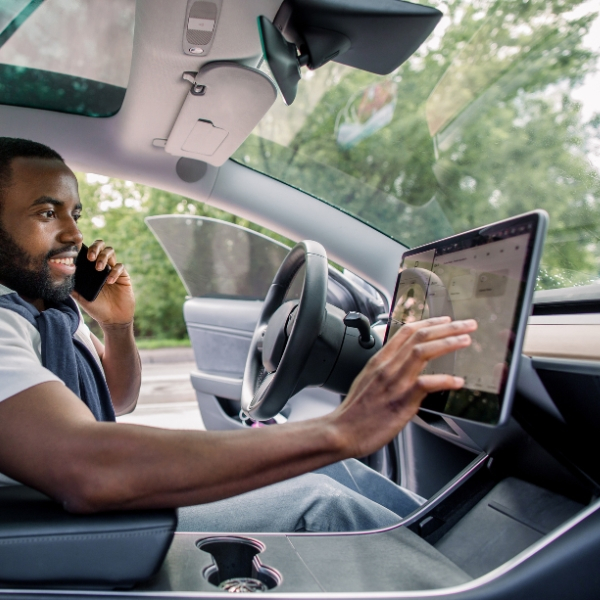 A man with a beard is sitting in the driver's seat of a car, talking on a smartphone with his left hand while touching the car's large central display with his right hand.
