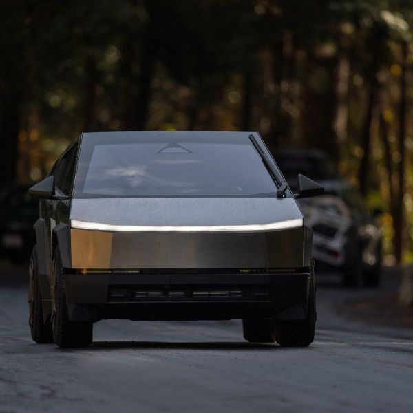A front view of a futuristic, angular dark grey vehicle with a prominent horizontal light bar across the front, driving on a road surrounded by trees.