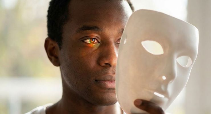 A person of color holding a white mask, revealing a thoughtful expression and expressive eyes underneath.