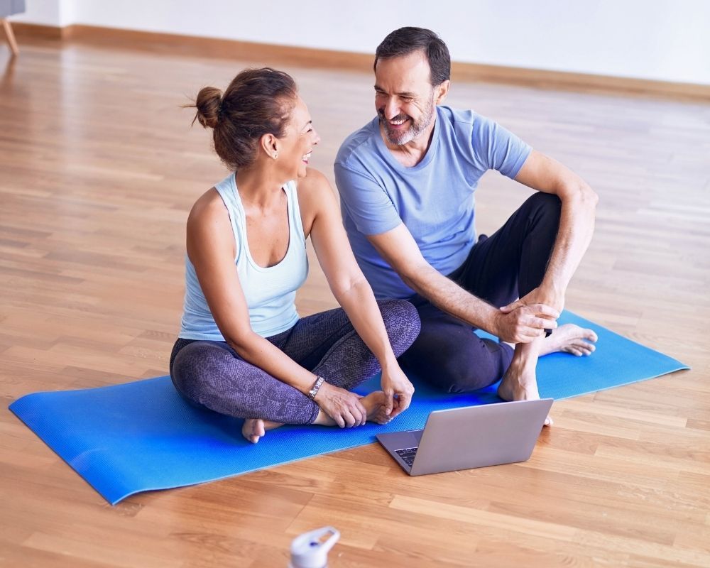 A diverse couple sitting cross-legged on a yoga mat in a studio, looking at a laptop, likely reviewing an online fitness class. A diverse couple sitting cross-legged on a yoga mat in a studio, looking at a laptop, likely reviewing an online fitness class.