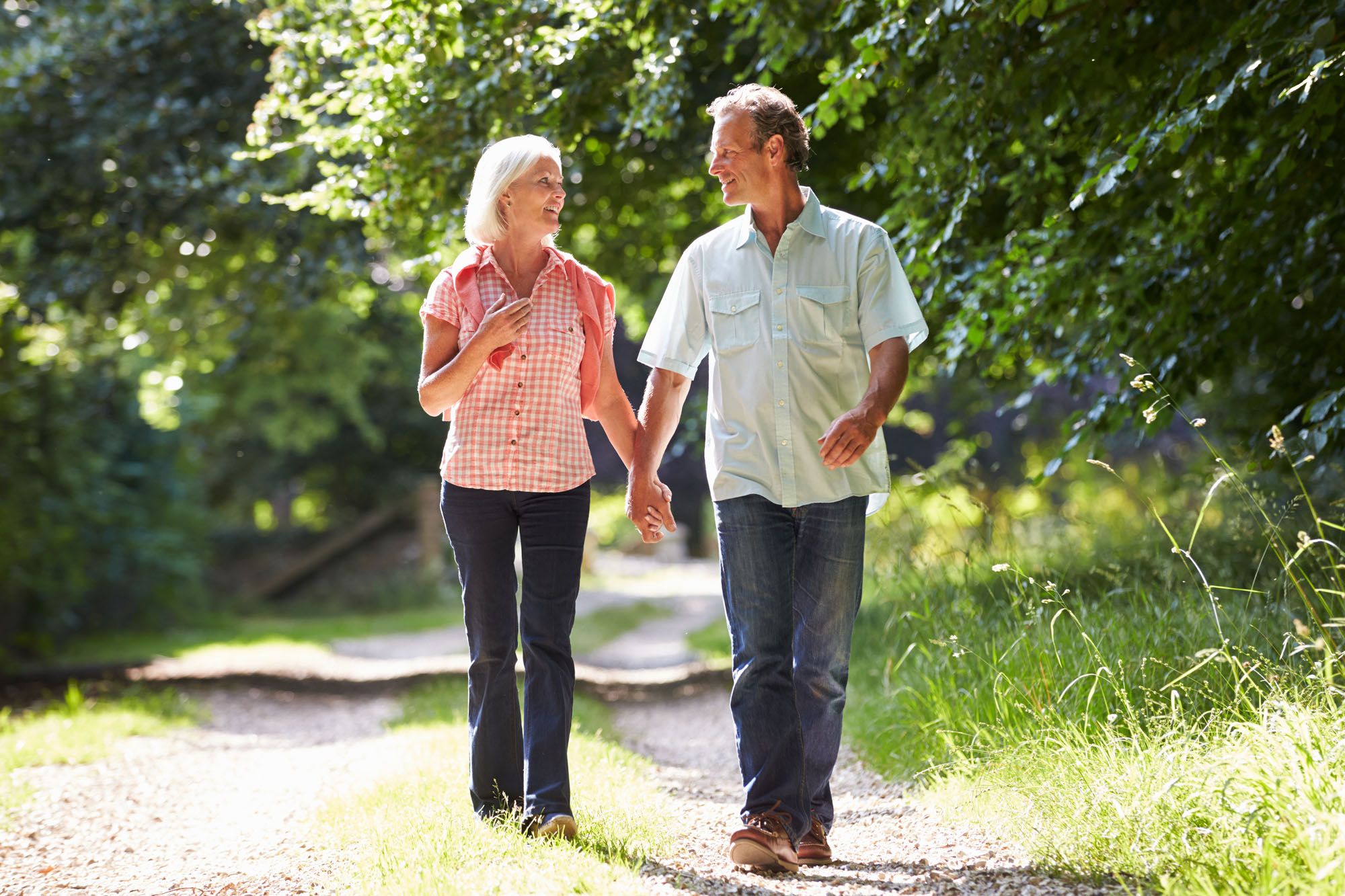 An older couple holding hands and smiling while walking down a sunlit dirt path bordered by trees. An older couple holding hands and smiling while walking down a sunlit dirt path bordered by trees.