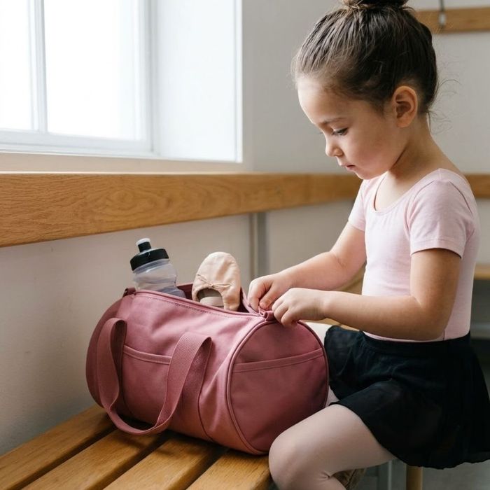 A young child sits on a wooden bench and zips up a durable pink duffel bag