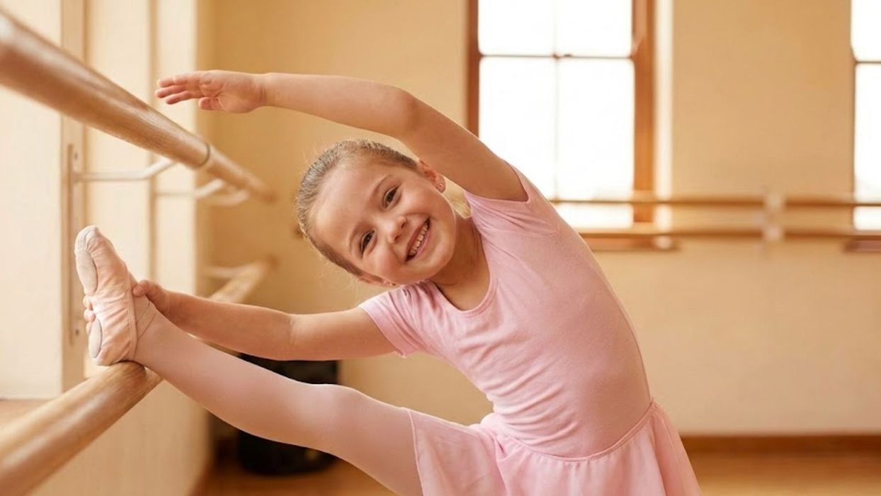 A young girl in pink dance attire smiles brightly while stretching at the barre.