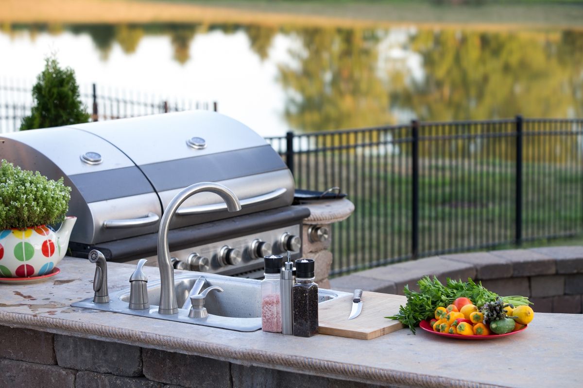 Preparing a healthy summer meal in an outdoor kitchen with gas barbecue and sink on a brick patio overlooking a tranquil lake with tree reflections
