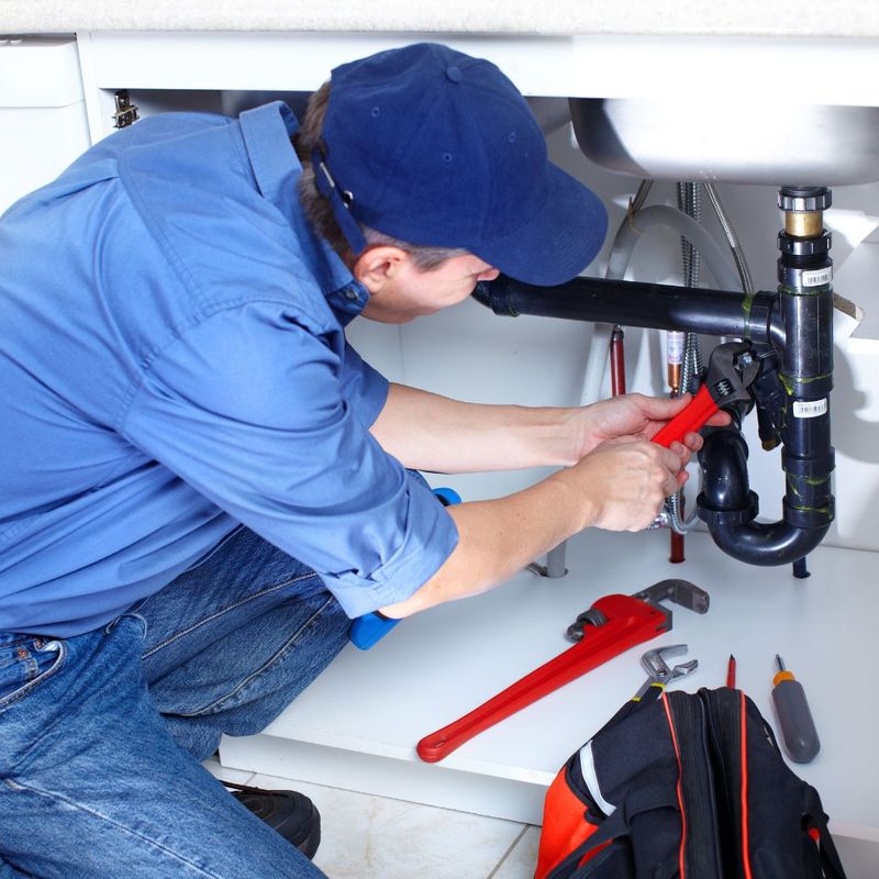 A person using a red pipe wrench to tighten a black plastic drainage pipe underneath a sink.
