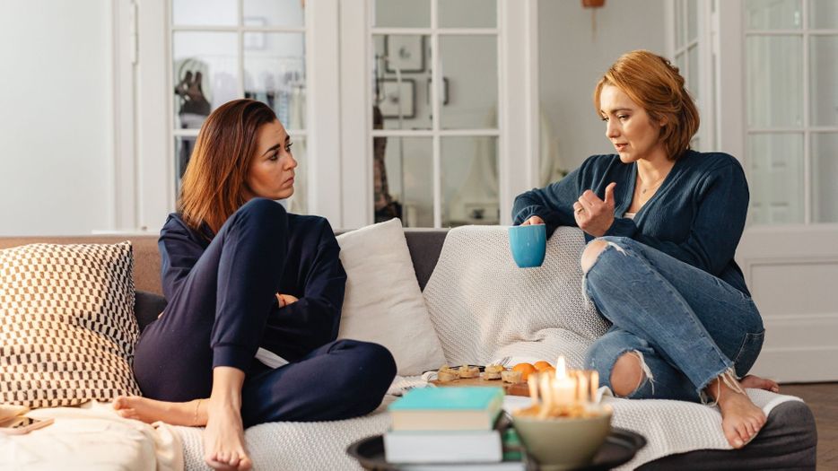 women sit on couch with coffee cups talking