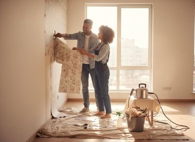 A happy couple smiles, working together to strip floral wallpaper from a wall in a bright room during home renovation. Couple Renovating Room