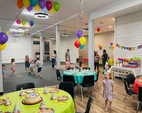 A vibrant kids' birthday party in a spacious venue, decorated with colorful ceiling balloons, streamers, and festive table settings.