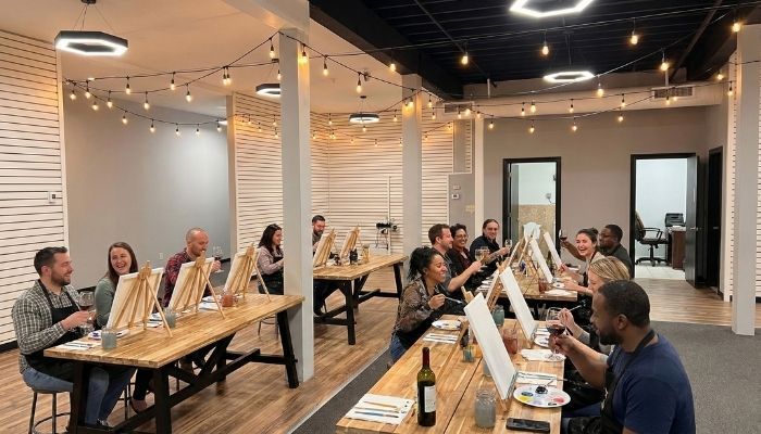 : Realistic photography of a "paint and sip" private event in a modern venue with guests painting at long wooden tables.