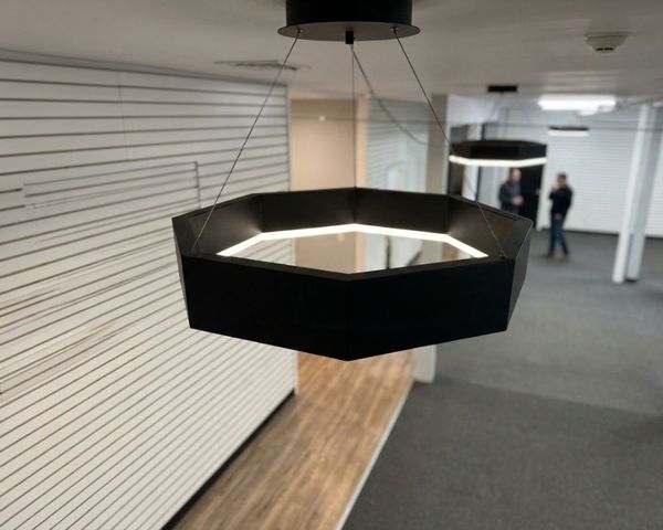 A contemporary architectural detail shot of a black hexagonal LED pendant light hanging from a high ceiling in a modern hall.