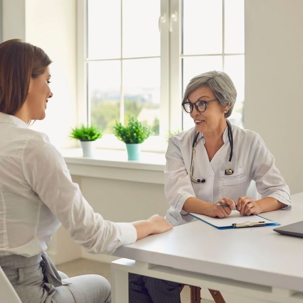 An experienced female doctor with grey hair and glasses, wearing a white lab coat and stethoscope, sits at a desk, smiling and actively listening to a female patient. The patient is seen from the back, in profile, dressed in a white shirt and 
