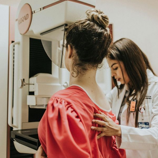 A woman in a red top stands facing a mammography machine, with her back to the viewer. A female medical professional in a white lab coat gently places a hand on the patient's back, guiding her during the breast screening procedure.