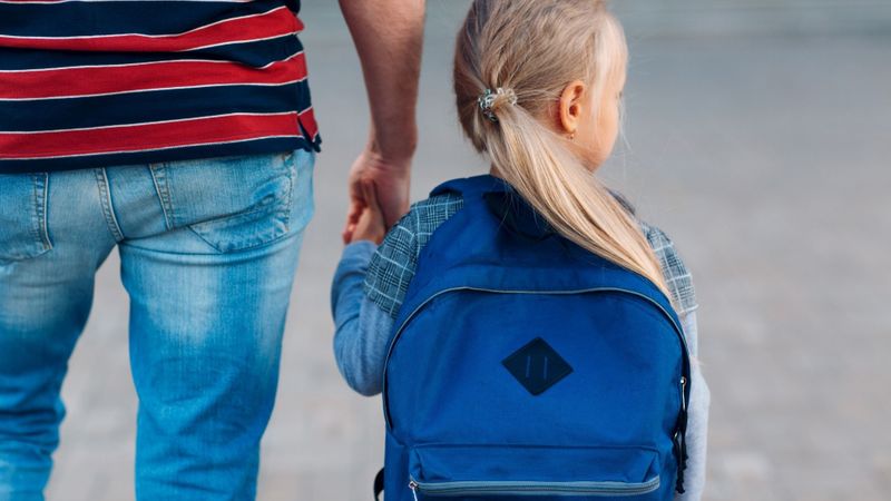 parent holds child's hand walking to school wearing a backpack