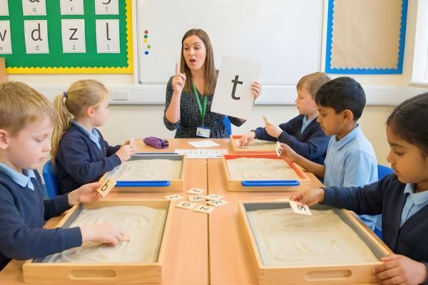 a female teacher leading a multi-sensory phonics lesson, holding up a letter card while small children trace letters in sand trays