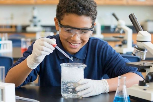 a young student with safety goggles, smiling and actively conducting a science experiment with a beaker and dropper in a lab